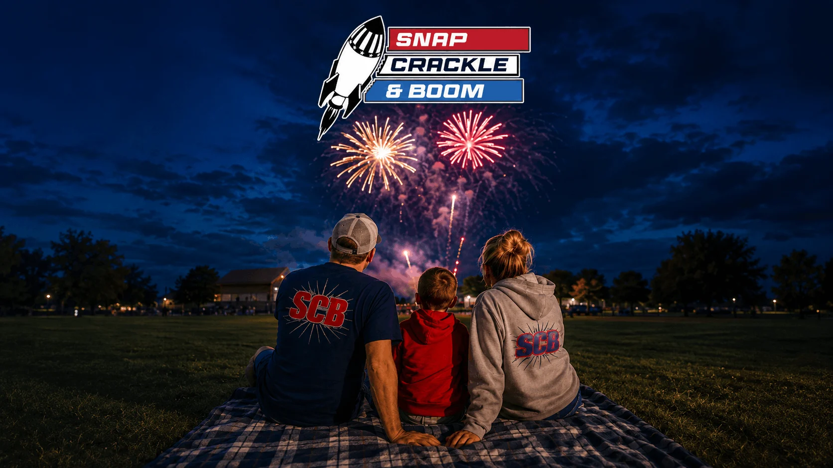 Family of three sits on a plaid blanket in a park at night, watching bright fireworks; adults wear SCB shirts and a child sits between them, with a 'SNAP CRACKLE & BOOM' banner overhead.